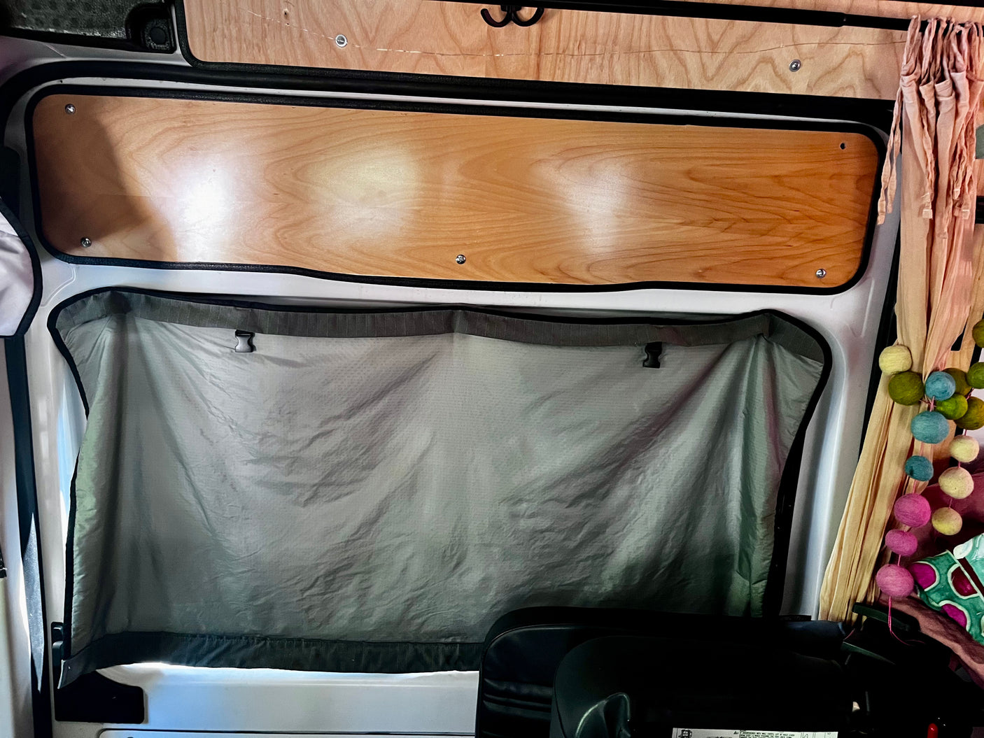 Interior of an RV rental, showing a window with a grey privacy shade, wooden paneling, and decorative felt balls for a cozy camper aesthetic.