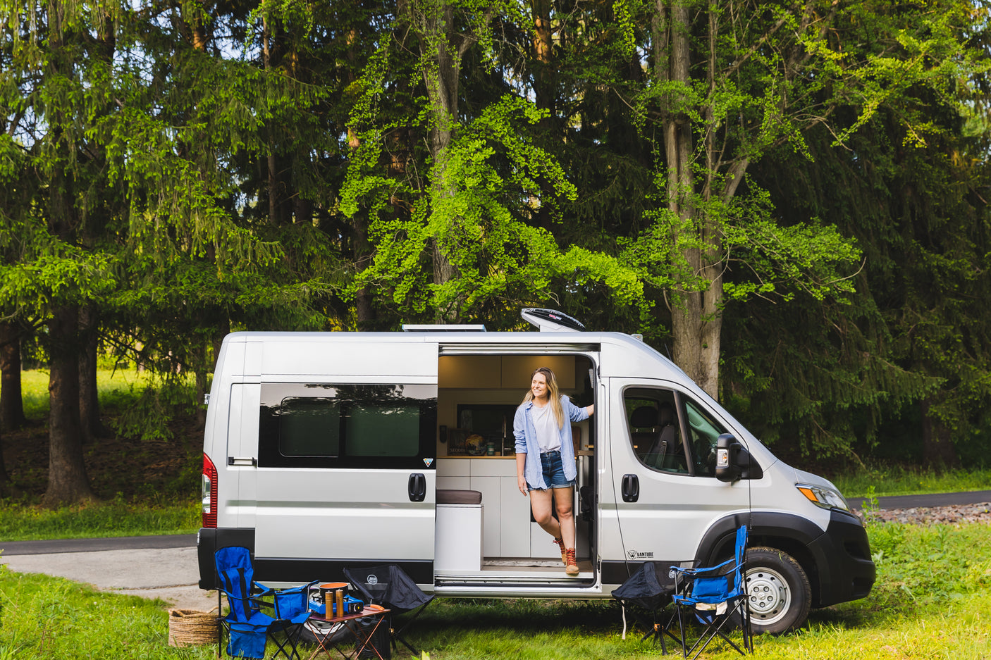 A smiling woman stands in her modern rv rental camper van, parked in a vibrant forest with camping gear ready.