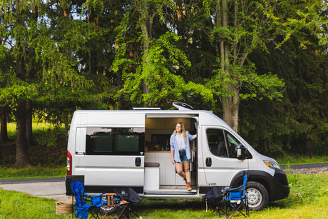 A smiling woman stands in her modern rv rental camper van, parked in a vibrant forest with camping gear ready.