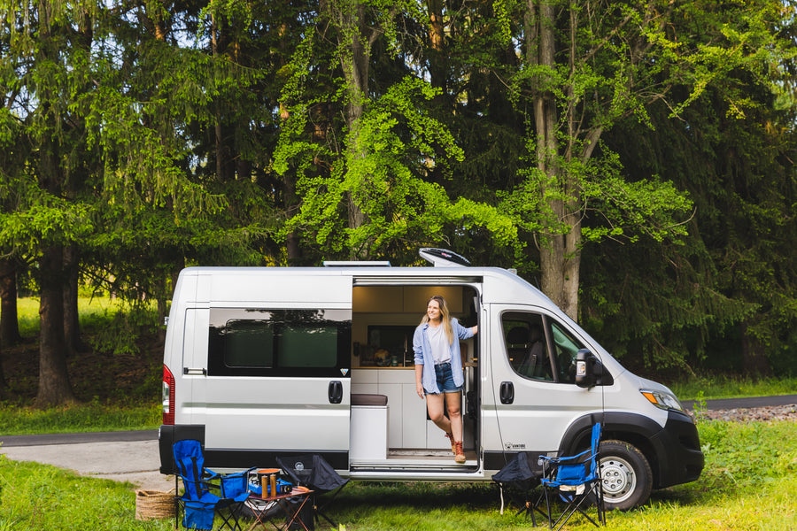 A smiling woman stands in her modern rv rental camper van, parked in a vibrant forest with camping gear ready.