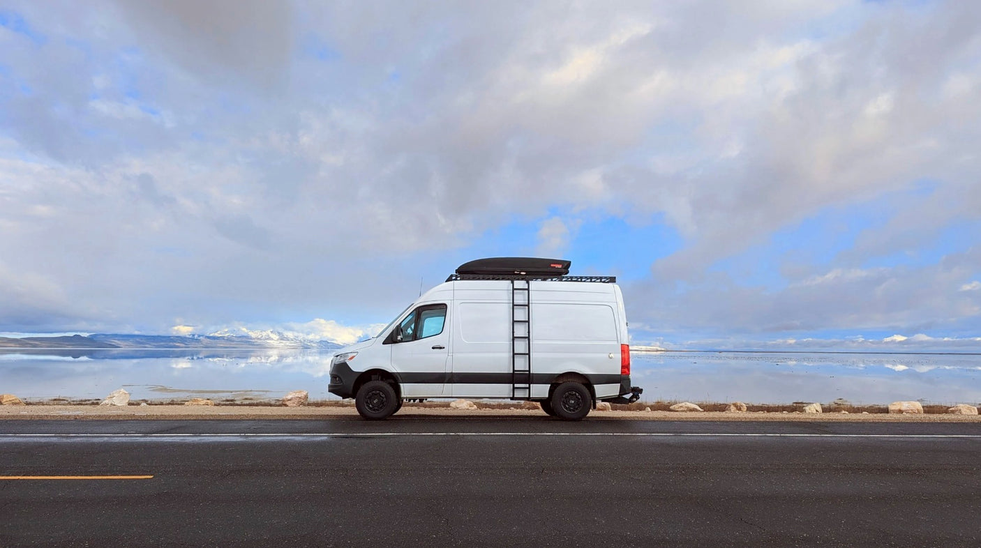White Sprinter RV rental parked on a road beside a vast, reflective lake. Snow-capped mountains and cloudy skies in background.