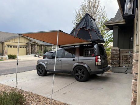 This gray SUV with a rooftop tent and awning could be an RV rental, parked in a suburban driveway.