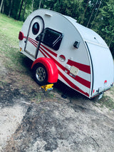 White T@G teardrop RV rental with red stripes, set up for camping in a wooded outdoor site. Perfect for travel adventures.