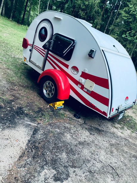 White T@G teardrop RV rental with red stripes, set up for camping in a wooded outdoor site. Perfect for travel adventures.