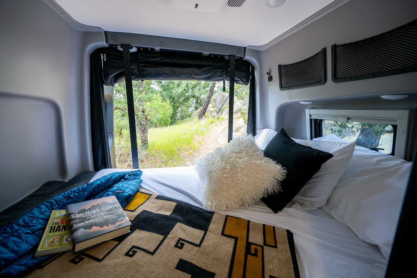 Cozy rv rental bedroom, featuring a large window with a scenic nature view, books, and blankets for comfort.