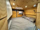 Warm RV rental bedroom with wood paneling, gray bedding, overhead lights, a roof vent, and wall-mounted controls. Left side storage.