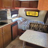 RV rental interior: kitchen sink, counter, cabinets, bed, and dinette with patterned cushions. Red LED lights.