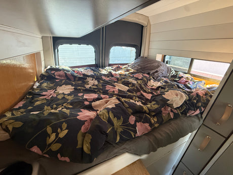 Floral duvet on a cozy bed inside an RV rental camper, with windows offering views outside.