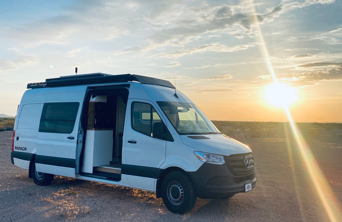 White Mercedes Sprinter camper van (RV rental) parked in desert at sunset, side door open. Ideal for road trip adventures.