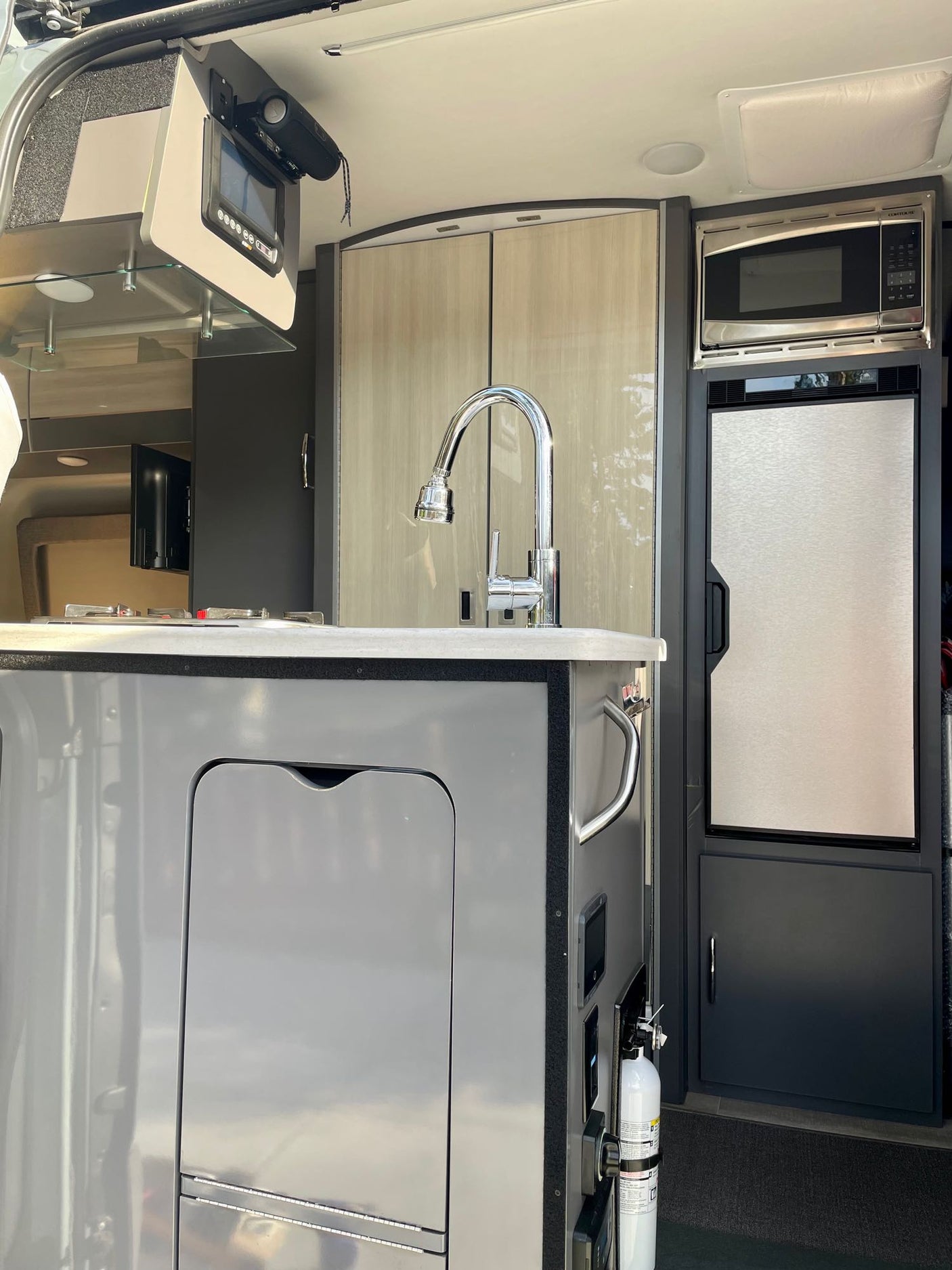 Compact RV rental kitchen with sink, faucet, microwave, and refrigerator visible, along with an overhead control screen.