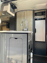 Compact RV rental kitchen with sink, faucet, microwave, and refrigerator visible, along with an overhead control screen.
