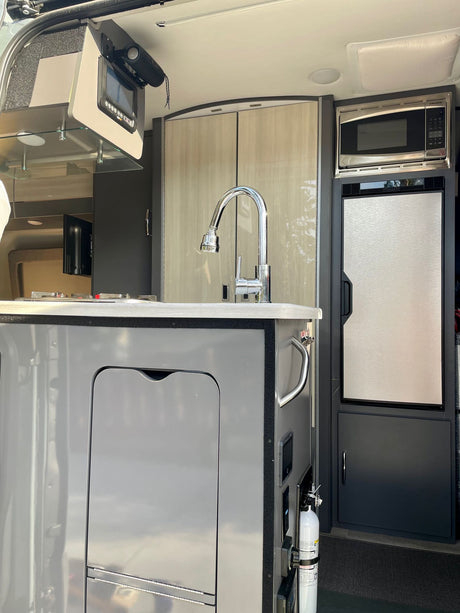 Compact RV rental kitchen with sink, faucet, microwave, and refrigerator visible, along with an overhead control screen.