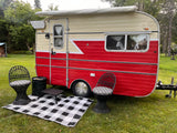 A charming vintage red & cream RV rental with a cozy outdoor seating area, including chairs & rug, set in a grassy natural space.