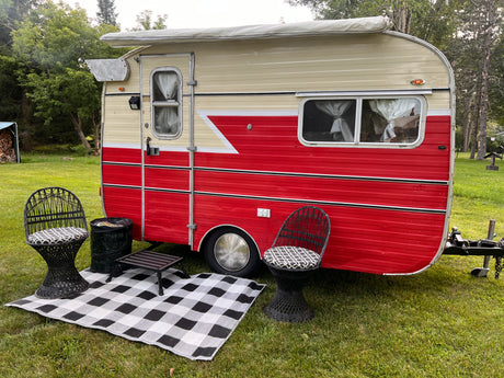 A charming vintage red & cream RV rental with a cozy outdoor seating area, including chairs & rug, set in a grassy natural space.