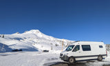 An RV rental van parked in a snow-covered lot at the base of a majestic, snow-capped mountain under a clear blue sky.