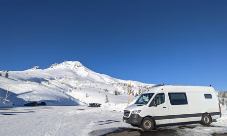 An RV rental van parked in a snow-covered lot at the base of a majestic, snow-capped mountain under a clear blue sky.
