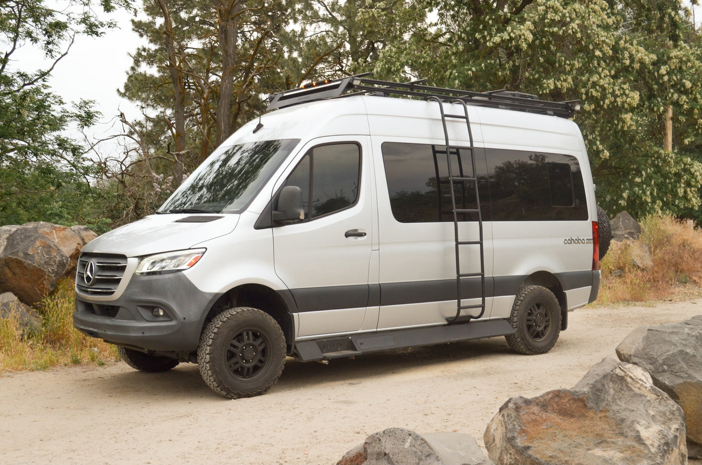 A white Mercedes-Benz Sprinter rv rental, equipped with a roof rack and ladder, parked on a dirt road by trees.