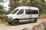 A white Mercedes-Benz Sprinter rv rental, equipped with a roof rack and ladder, parked on a dirt road by trees.