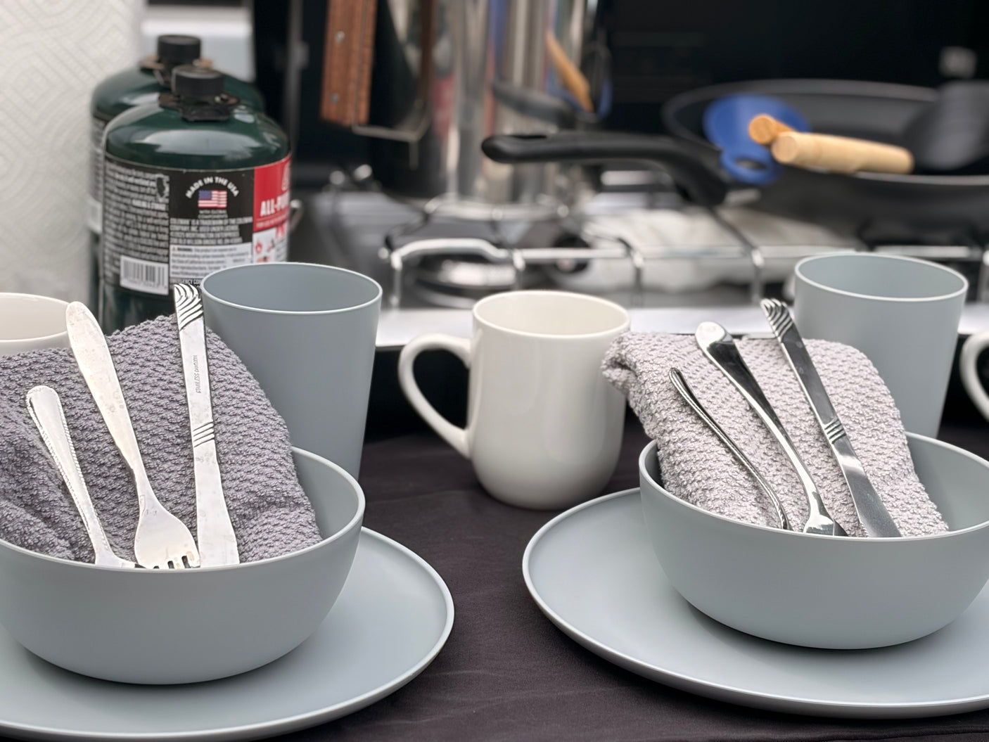 Camping dinnerware, complete with silverware and towels, alongside an outdoor cooking setup with propane tanks and a stove, ready for your RV rental adventure.