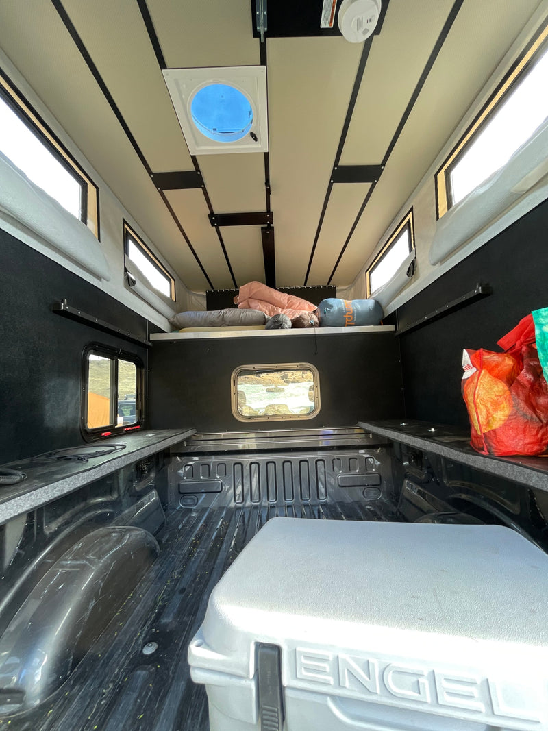 Inside an RV rental truck bed camper: A cozy sleeping platform, side storage, Engel cooler, windows, and roof vent are visible. — photo 10