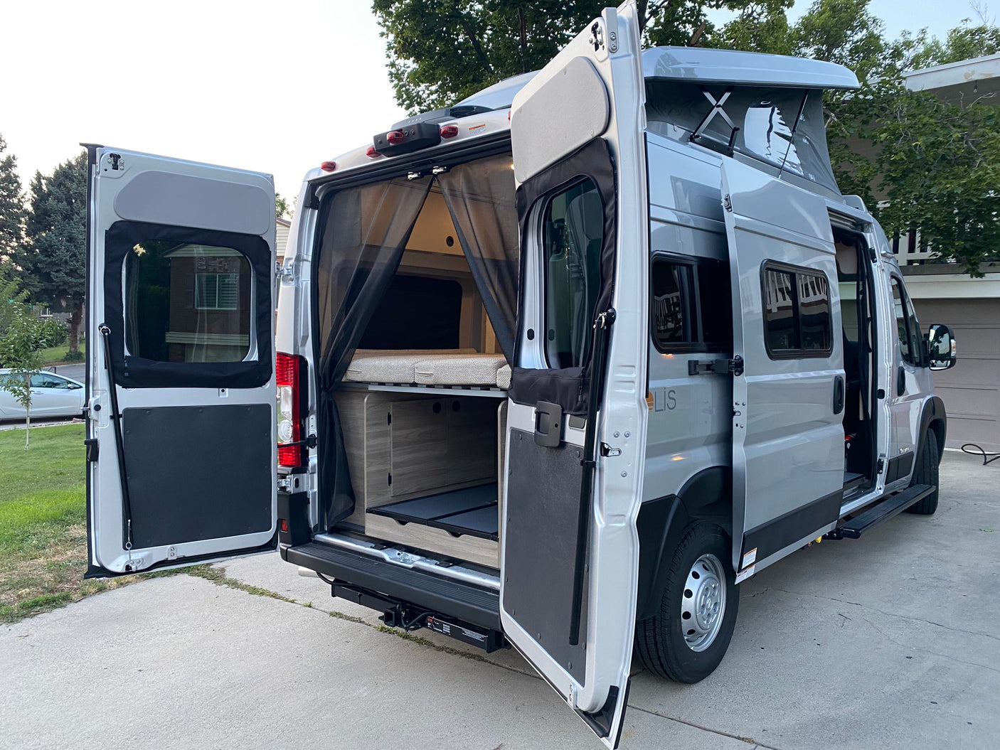 This silver RV rental camper van has its rear and side doors open, showcasing the interior bed, storage, and raised pop-top roof.