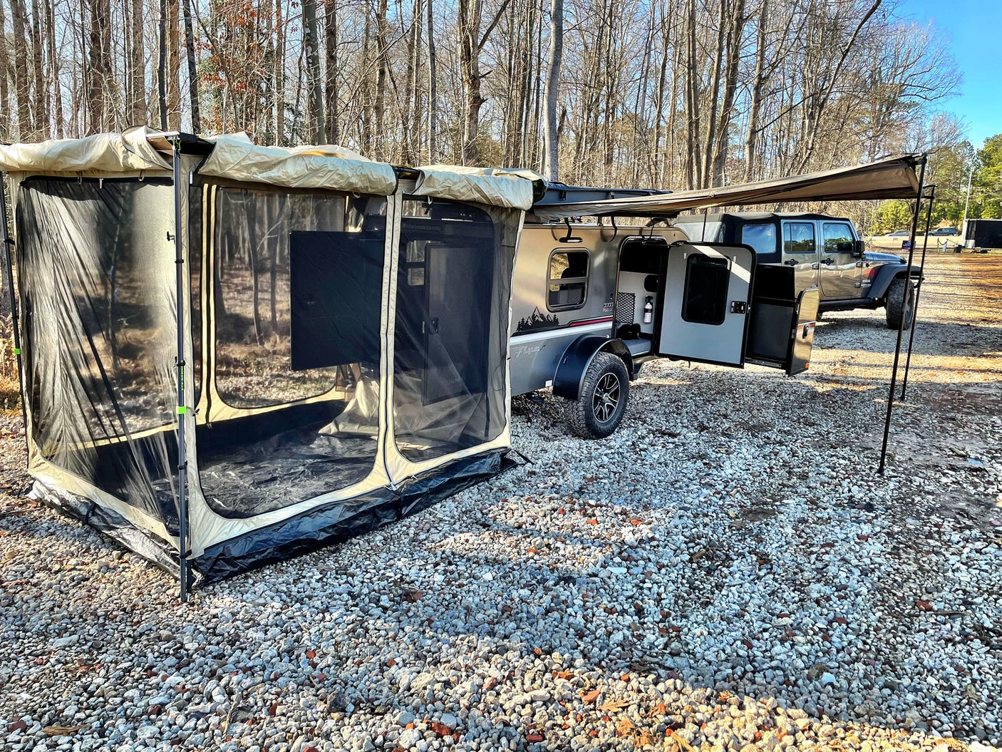 Compact rv rental: a teardrop camper trailer with an attached screen room and awning, on gravel, by a Jeep.