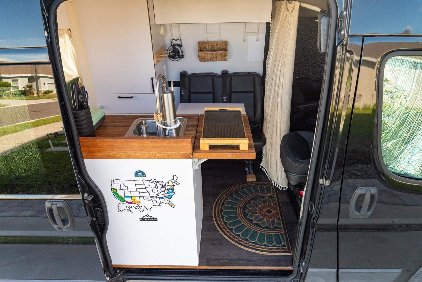 RV rental interior: a compact kitchen with wood counters, sink, portable grill, black seating, & decorative floor mat. — photo 5
