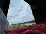 An RV rental's comfy red bed blanket offers a scenic view of a calm lake, green hills, and cloudy sky.