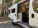 Discover freedom with this RV rental. White camper van in a sunny forest, its side door open to reveal a cozy interior and pull-out table.