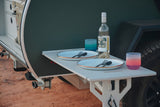An RV rental's clever pull-out table is set for two with plates, cutlery, a wine bottle, and colorful glasses for outdoor meals.