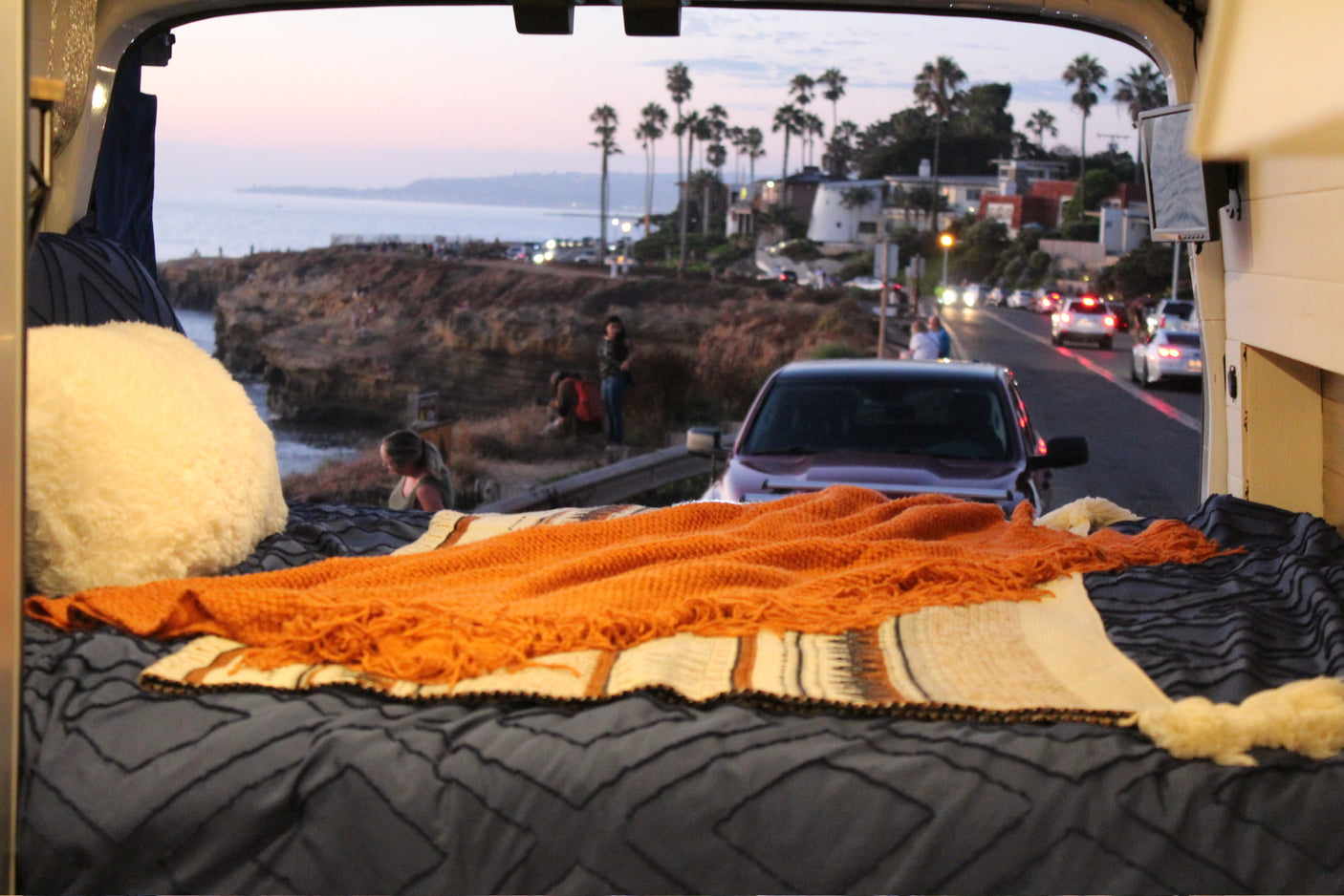 Inside a cozy RV rental, a comfy bed faces a scenic coastal view with cliffs, ocean, palm trees, and road.