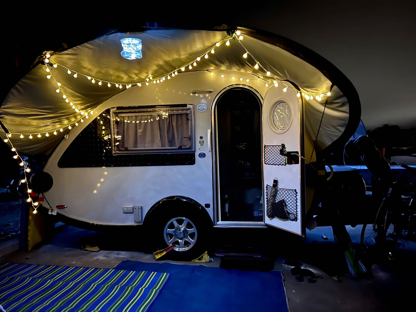 Experience a cozy night: This RV rental teardrop trailer is illuminated by string lights under its awning for camping.