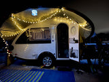 Experience a cozy night: This RV rental teardrop trailer is illuminated by string lights under its awning for camping.
