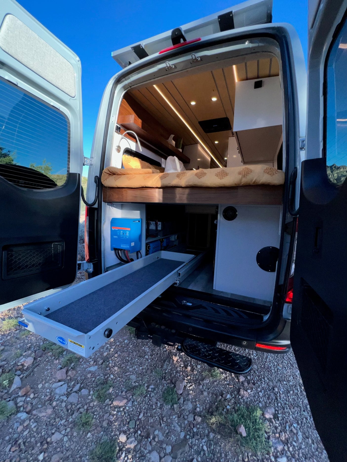 Inside an RV rental, open back doors reveal a camper van's cozy bed, pull-out storage, and Victron power setup.