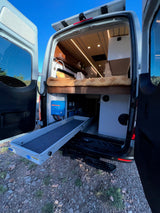 Inside an RV rental, open back doors reveal a camper van's cozy bed, pull-out storage, and Victron power setup.