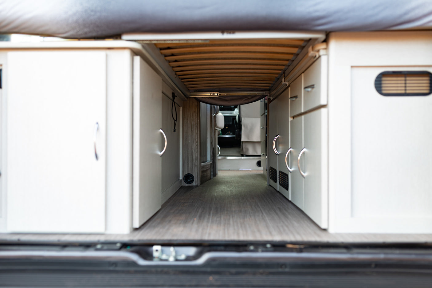 Inside a modern **rv rental**, looking from the rear. Features a raised bed platform, wood floor, and sleek white storage cabinets.