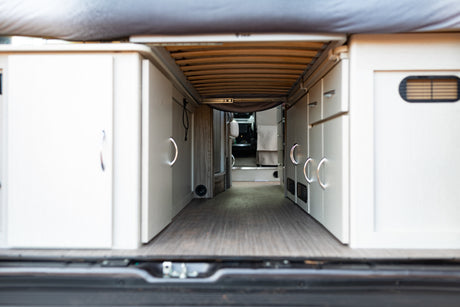 Inside a modern **rv rental**, looking from the rear. Features a raised bed platform, wood floor, and sleek white storage cabinets.
