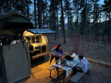 Dusk at a forest RV rental campsite, with a camper trailer & rooftop tent, where two people are cooking.