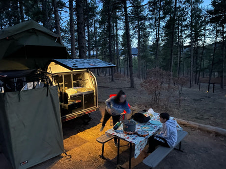 Dusk at a forest RV rental campsite, with a camper trailer & rooftop tent, where two people are cooking.