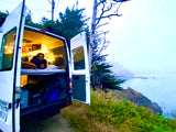 Cozy rv rental experience: A man relaxing inside his campervan, parked overlooking a misty coastal ocean view.