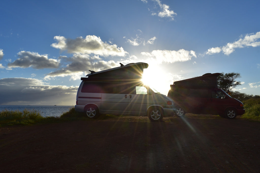Silhouetted RV rentals, two camper vans (one silver, one red with surfboard) parked by the ocean at sunset.