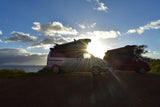Silhouetted RV rentals, two camper vans (one silver, one red with surfboard) parked by the ocean at sunset.