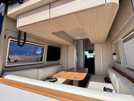 Enjoy this bright RV rental interior featuring light wood, cream seating, a versatile table, and a scenic beach view from the window.