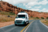 A white Mercedes-Benz Sprinter van, perfect for an rv rental adventure, drives past towering red rock cliffs on a scenic road.