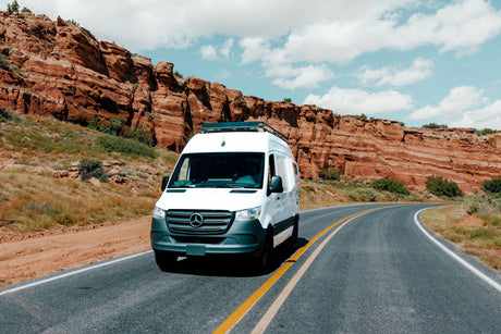 A white Mercedes-Benz Sprinter van, perfect for an rv rental adventure, drives past towering red rock cliffs on a scenic road.