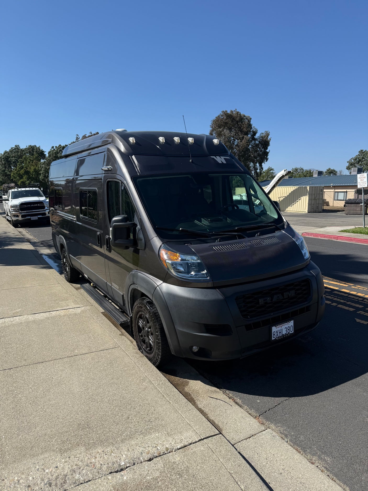A dark gray Ram ProMaster camper van with rooftop lights, ideal for an rv rental, parked on a sunny street.