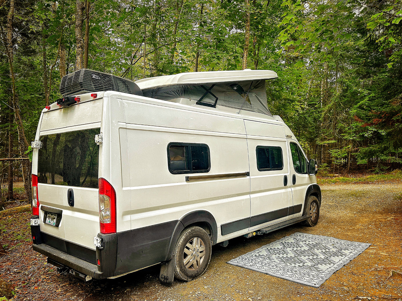 White pop-top camper van RV rental parked in a tranquil forest setting with an outdoor mat.