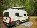 White pop-top camper van RV rental parked in a tranquil forest setting with an outdoor mat.