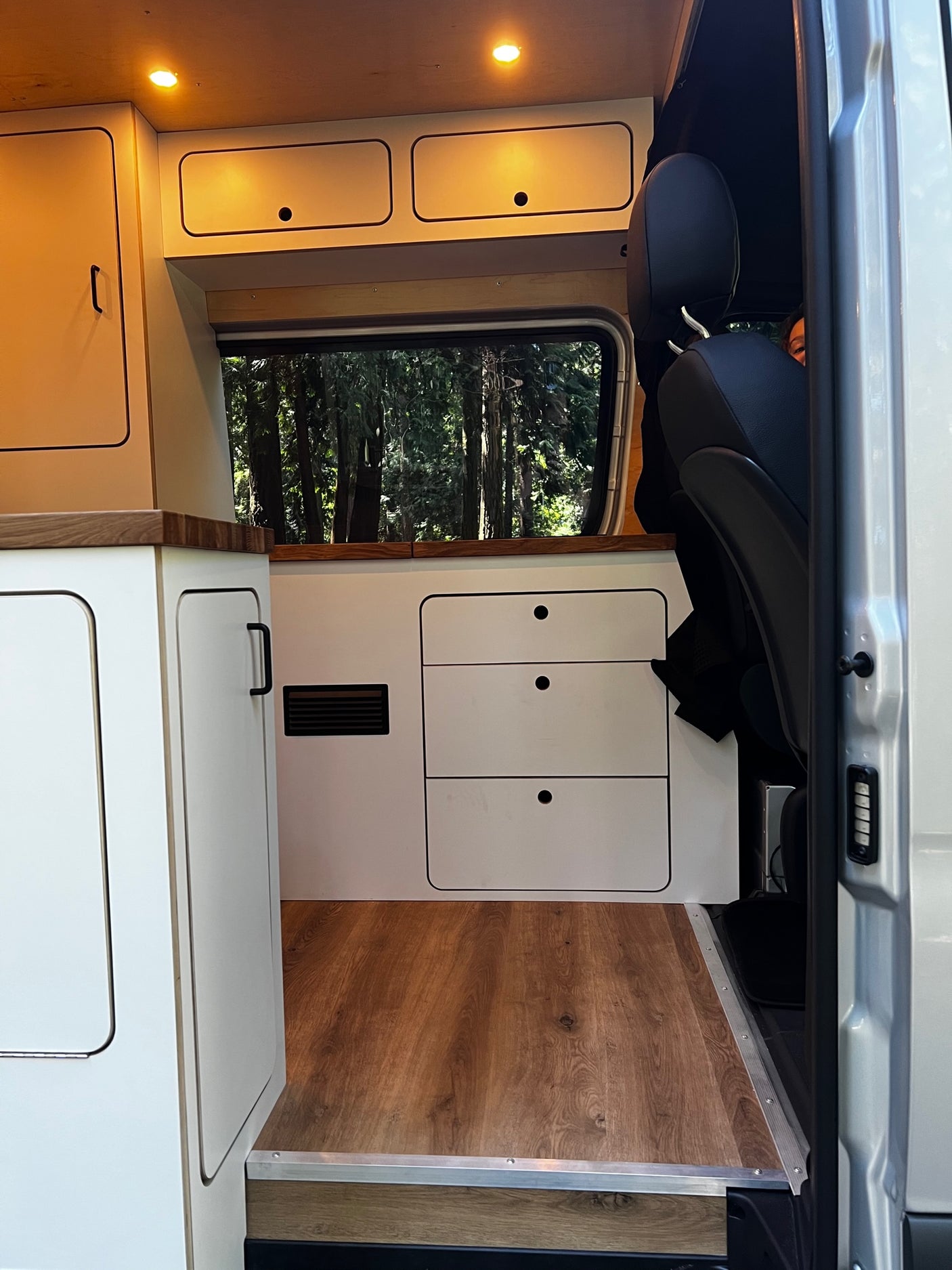 Modern RV rental interior: Clean white cabinets, wood paneling, ambient lighting, and a window view of trees.