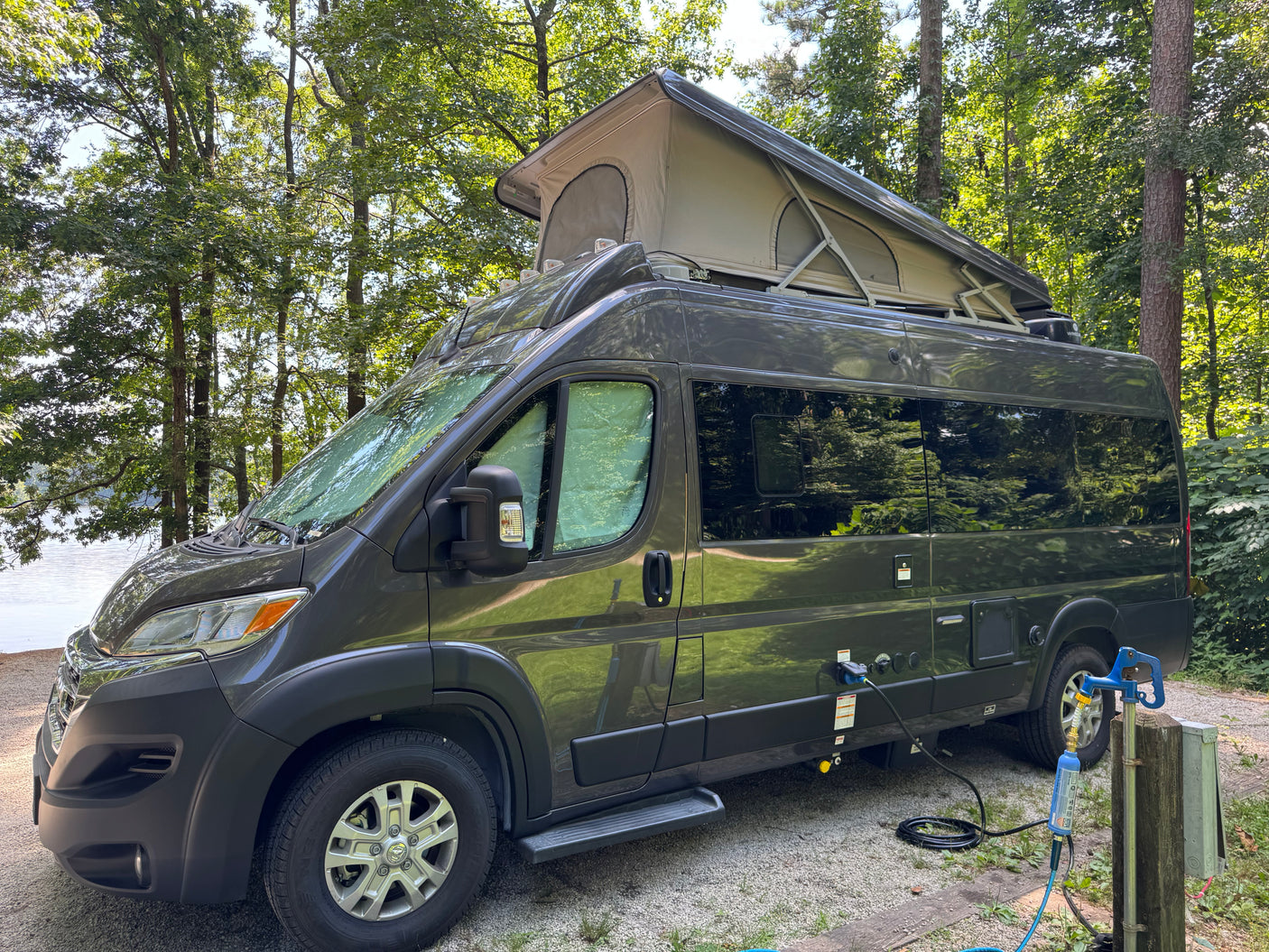 Modern gray rv rental camper van with a pop-top roof, parked at a scenic lakeside campsite and hooked up to utilities.